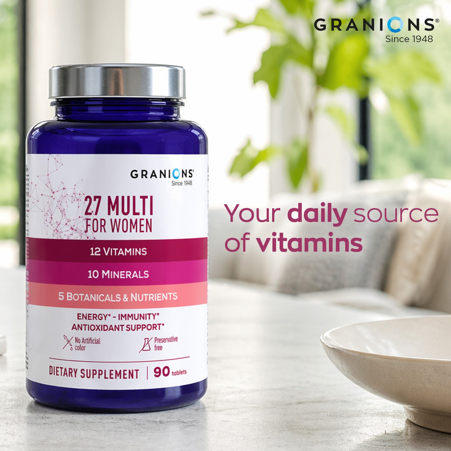 A bottle of Granicns 27 Multi for Women dietary supplement placed on a marble countertop with a blurred living room background and a bowl of food nearby. The bottle is dark blue with a silver cap and a white label featuring pink and magenta accents.